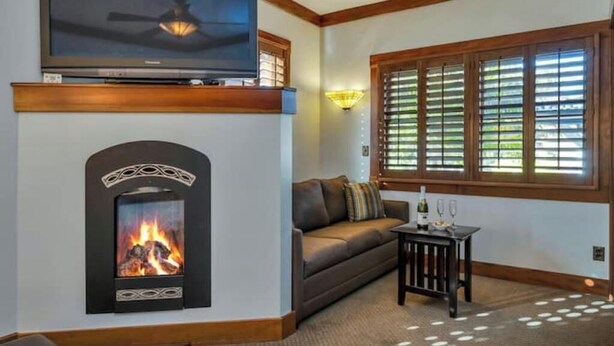 A cozy living room with a fireplace, a flat-screen TV mounted above it, a comfortable sofa, and a small table with glasses and a bottle.