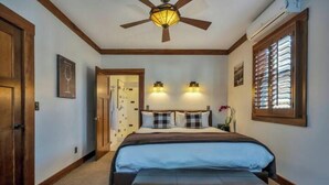 A bedroom with a bed, a ceiling fan, two wall-mounted lights, a window with blinds, and a door.