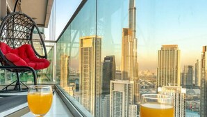 Un balcon avec une table en verre, une chaise rouge, un petit-déjeuner composé de croissants, de fraises et d’un verre de jus d’orange, ainsi qu’une bière, le tout offrant une vue sur un paysage urbain avec de hauts immeubles et le Burj Khalifa.