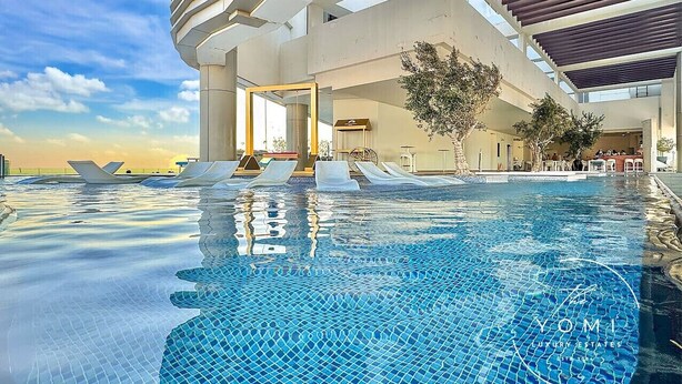 Une piscine à débordement moderne offrant une vue dégagée sur le ciel, un espace couvert avec des sièges et l’entrée du bâtiment.