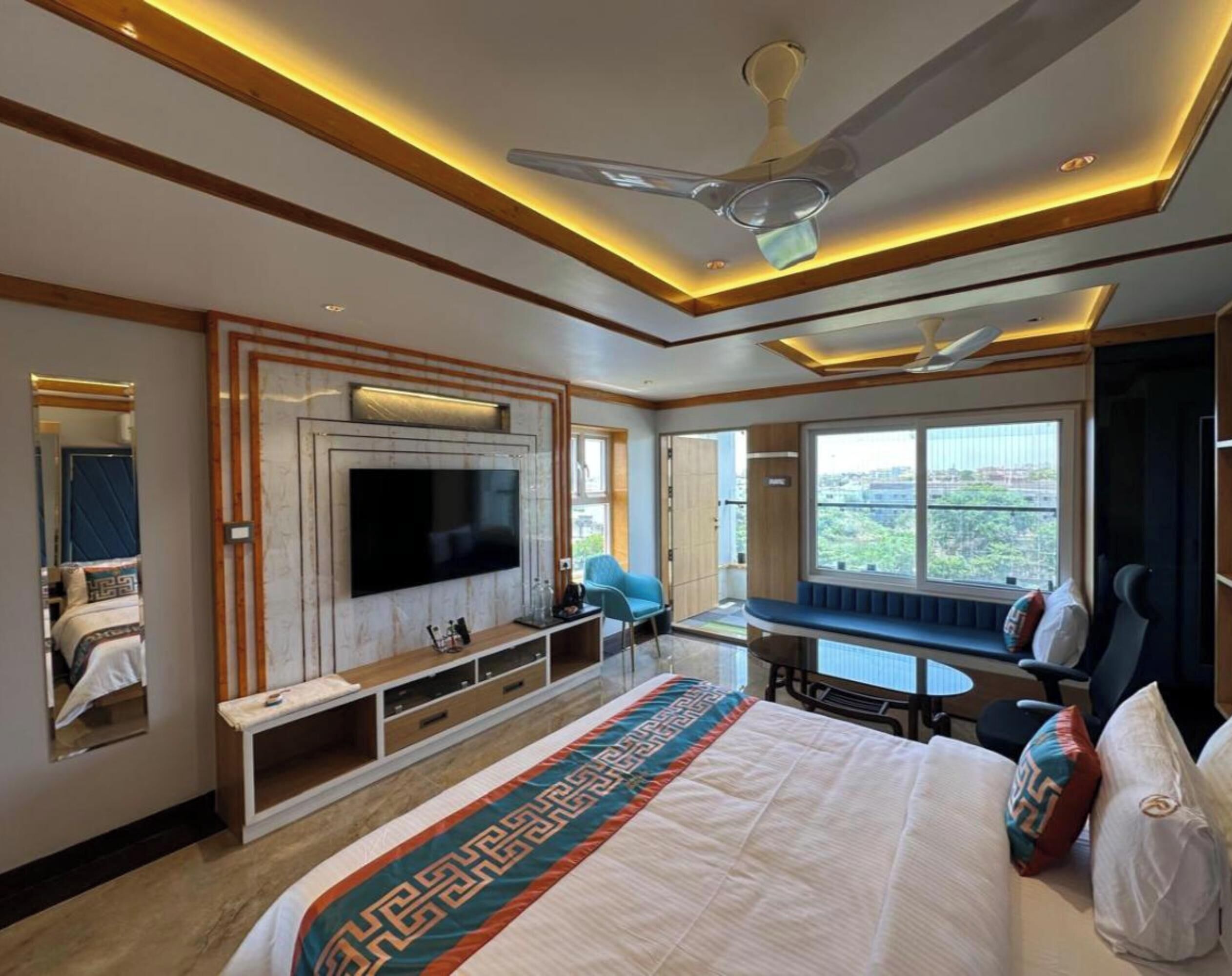 A modern hotel room with a large bed, a flat-screen TV, a seating area with a table, and a large window offering a city view.