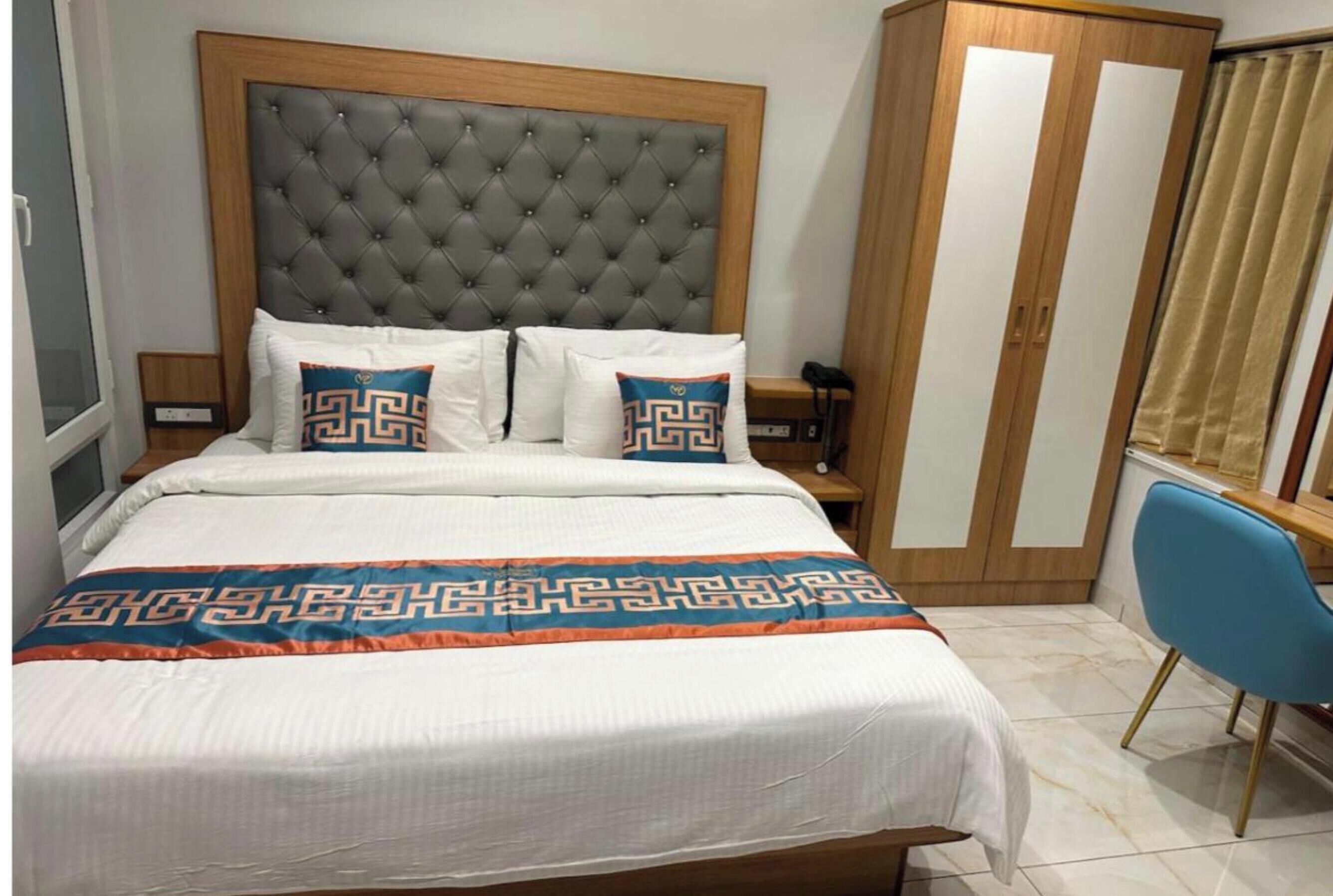 A bedroom with a tufted headboard, a bed with patterned bedding, a wardrobe, a desk, and a chair.