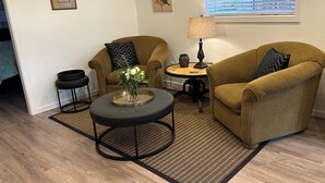 A living room with two armchairs, a coffee table, a lamp, and a small side table. There are framed pictures on the wall and a window with blinds. - Private suite steps away from the ferry and some of Powell River's best dining. (Powell River)
