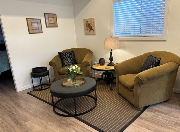 A living room with two armchairs, a coffee table, a lamp, and a small side table. There are framed pictures on the wall and a window with blinds.