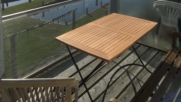 Un balcon avec une table et des chaises en bois, donnant sur une étendue d’eau calme et une côte lointaine.