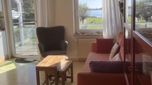 Un salon comprenant un canapé, un fauteuil et une table basse en bois. De grandes fenêtres, équipées de rideaux, laissent entrer la lumière, et on aperçoit un radiateur ainsi qu’une vue sur un lac.
