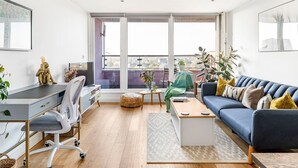 A modern living room with a blue sofa, a desk with a chair, a coffee table, and a view through large windows.
