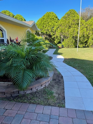 A pathway leading to a building with a yellow facade, surrounded by lush greenery and trees. - Cozy 2 Bedroom Sebring Garden Retreat ~ Private Entrance ~ AC and Wi-Fi (Sebring)