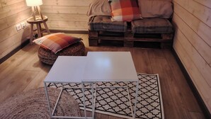 Ein gemütliches Zimmer mit Holzboden, einem weißen Tisch, einem gemusterten Teppich, einer Couch mit Kissen und einer Lampe auf einem kleinen Beistelltisch.