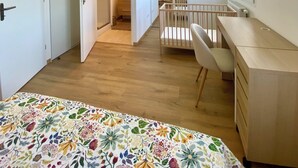 A bedroom with a bed, a desk, and a crib. There is a window and a door leading to a bathroom.