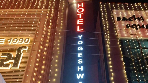 Ein Hotelschild mit der Aufschrift 'HOTEL YOGESHWAR ROOMS' und darunter der Schriftzug 'Zimmer rund um die Uhr geöffnet'.