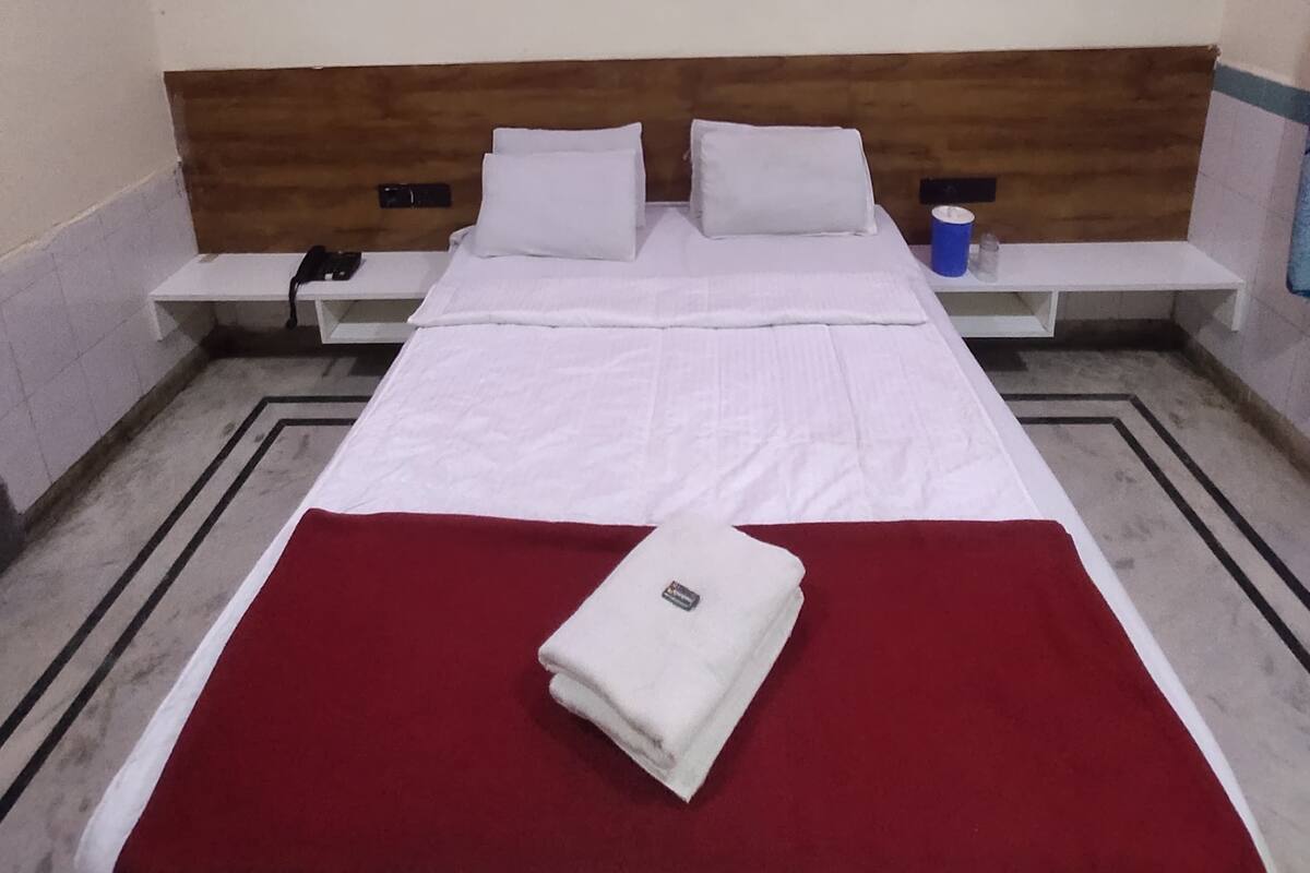 Quarto de hotel com cama, manta vermelha, ventoinha no teto e mesa de cabeceira com telefone e um recipiente azul.