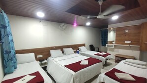 Ein Hotelzimmer mit vier Betten, jeweils mit weißer Bettwäsche und roten Decken. Es gibt einen Deckenventilator, einen an der Wand montierten Fernseher und einen kleinen Tisch mit einem Stuhl.