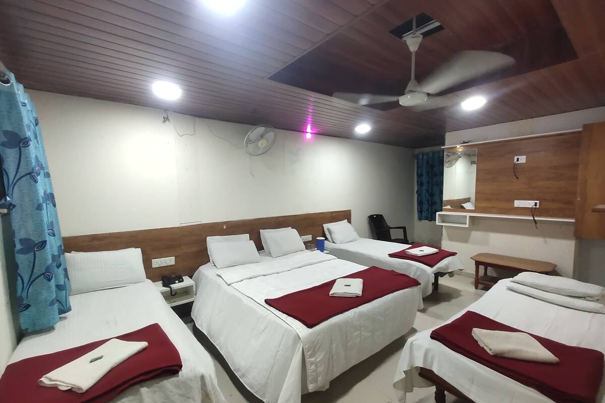 Quarto de hotel com quatro camas, cada uma com roupa de cama branca e cobertores vermelhos. Há um ventilador de teto, uma televisão fixada na parede e uma pequena mesa com uma cadeira.