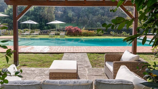 Un espacio de estar techado al aire libre con mobiliario de mimbre y sombrillas, con vistas a una piscina y un jardín exuberante.