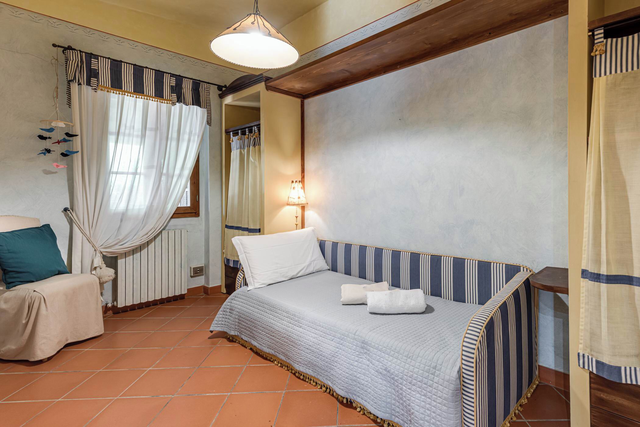 Quarto com cama, cadeira, luminária pendente e janela com cortinas.