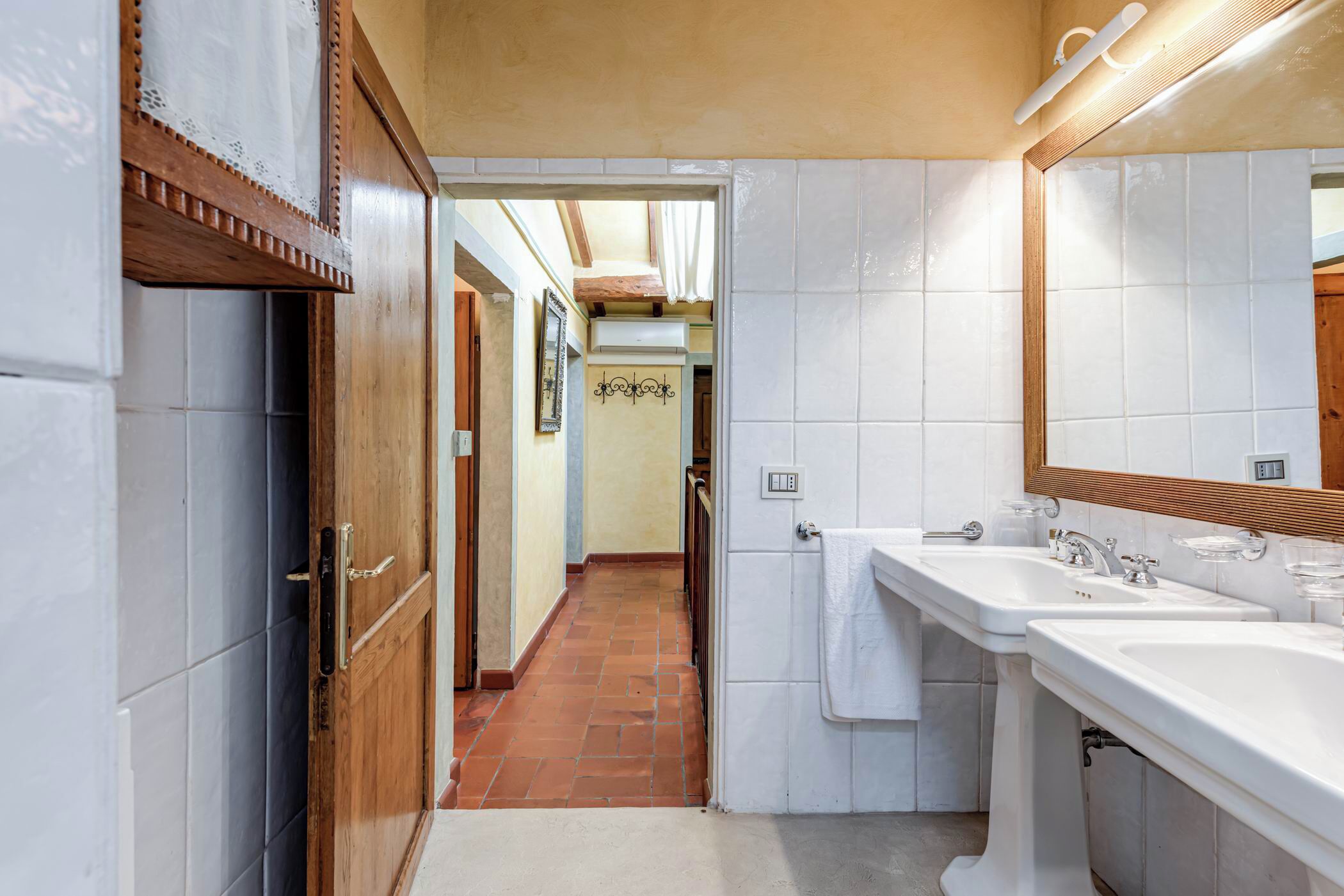 A narrow hallway with a wooden door, white tiled walls, and a tiled floor. There are two sinks with mirrors and a towel hanging on the wall.