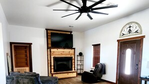 A living room with a wooden floor, a ceiling fan, a fireplace, and a television mounted above it. - Come enjoy the breathtaking views from the High Country Retreat (Mead)