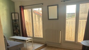A spacious room with a wooden floor, a sofa, a small table, and a radiator. There are two windows with a view of a fence and buildings outside.