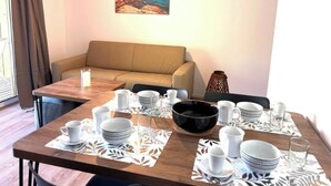 Un coin repas avec une table en bois dressée pour quatre personnes, un canapé beige et un tableau représentant un paysage côtier accroché au mur.