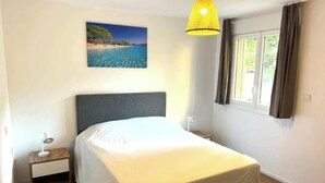 Une chambre à coucher avec un lit, une table de chevet, une lampe, une fenêtre avec des rideaux et un tableau représentant une scène de plage.