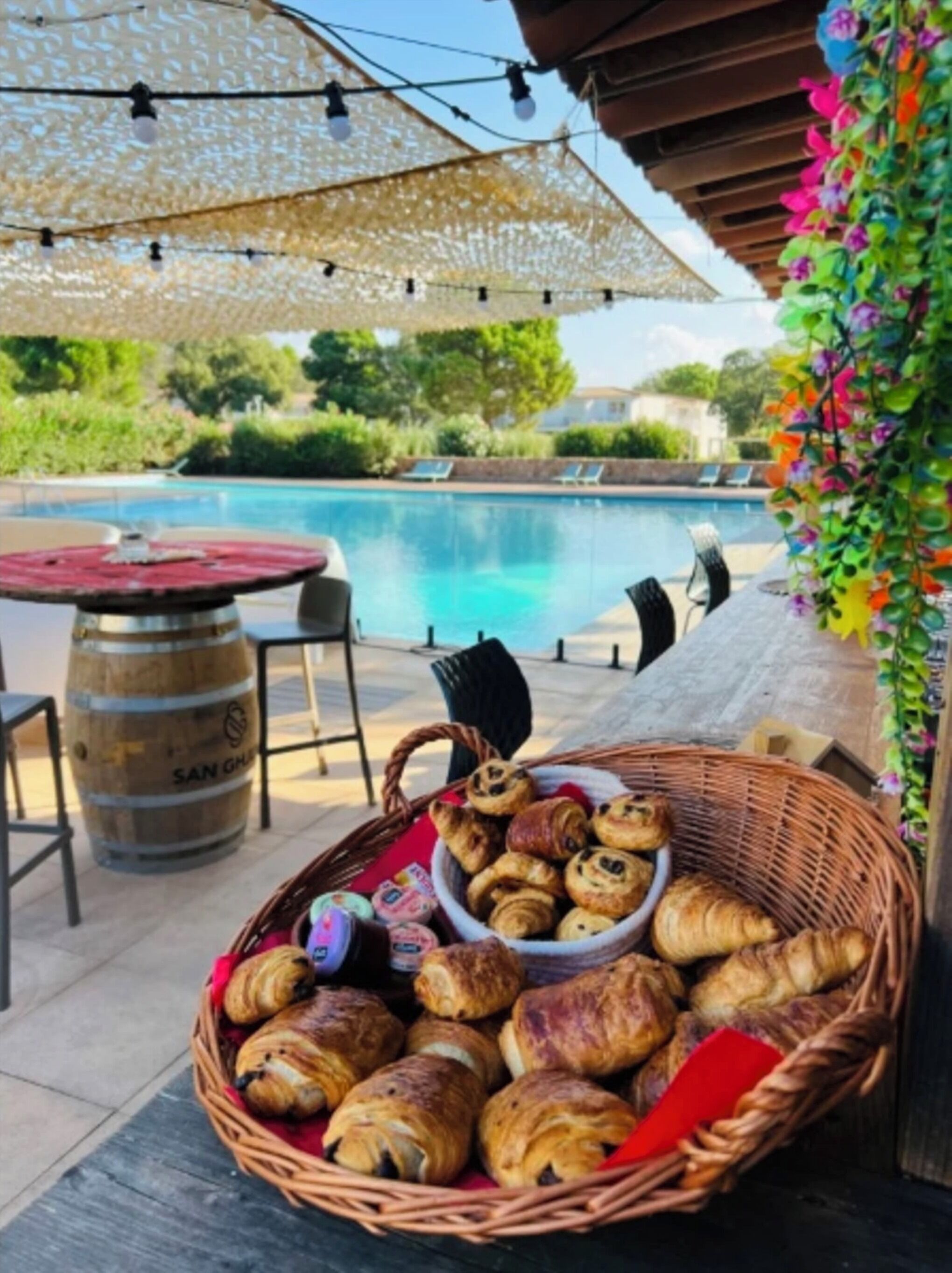 A poolside setting with a wicker basket of pastries, a wooden barrel, and outdoor seating.