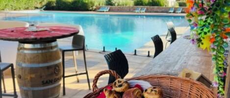 A poolside setting with a wicker basket of pastries, a wooden barrel, and outdoor seating.