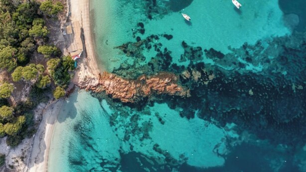 Vue aérienne d’une zone côtière avec des eaux turquoise limpides, des affleurements rocheux et une plage de sable.