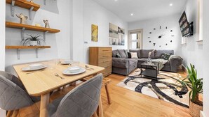 A modern living room with a dining area, grey sofas, wooden shelves, and a clock on the wall. - Modern Spacious 1 Bed Flat near Richmond Park and River Thames  (London)
