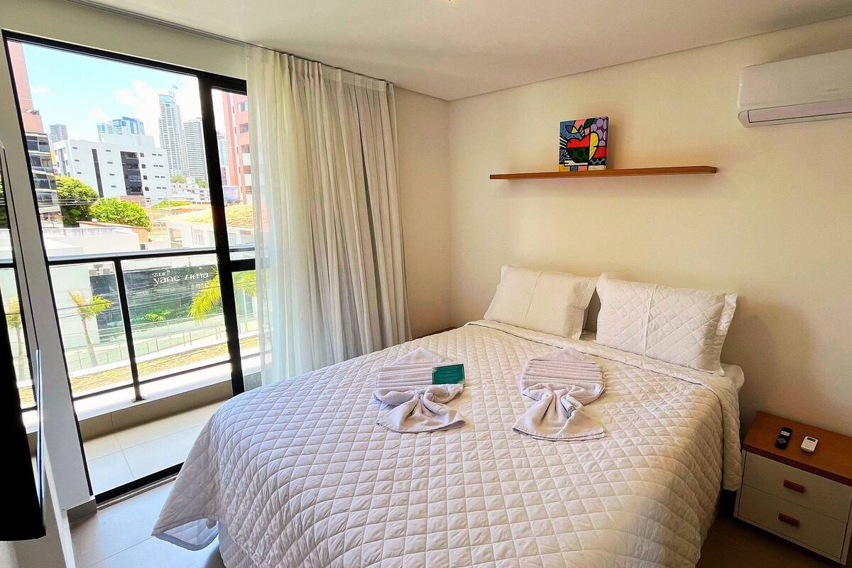 Une chambre d’hôtel avec une grande fenêtre, un lit avec une literie blanche, une petite table de chevet et un appareil de climatisation fixé au mur.