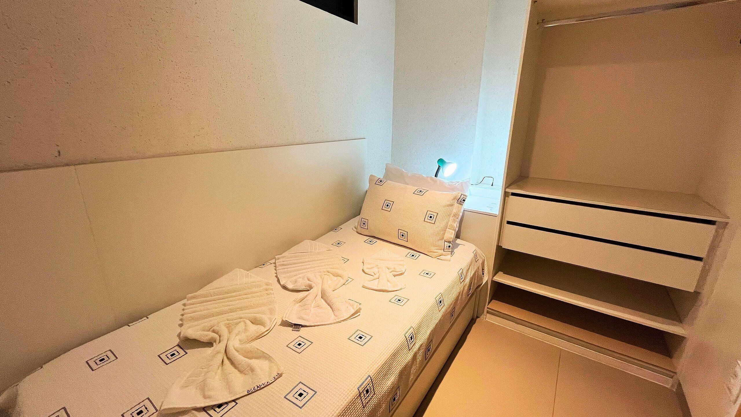 A small, neatly made bed with a patterned bedspread and pillows, a bedside table with a lamp, and a built-in shelf unit.