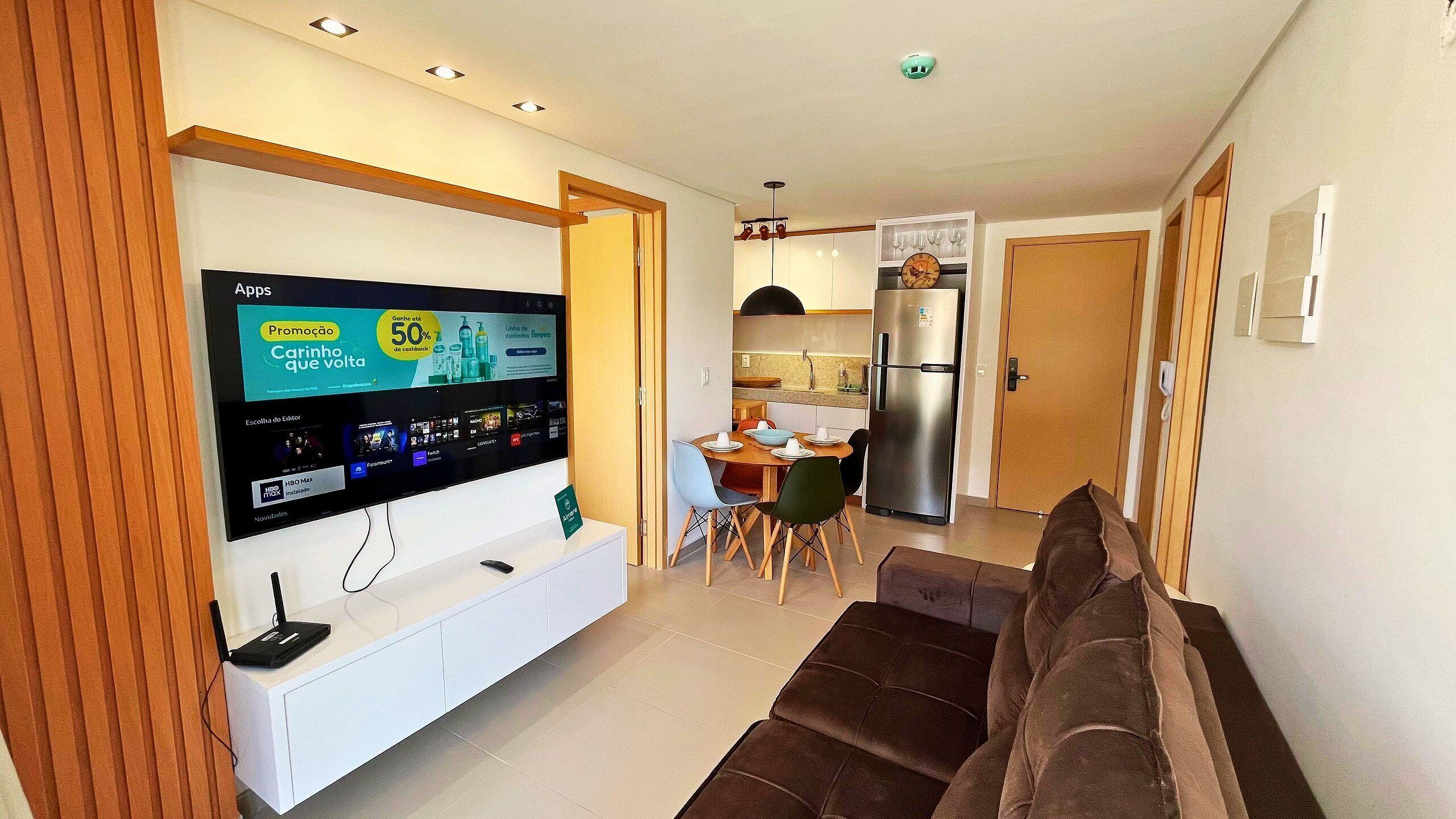 A modern living room with a flat-screen TV mounted on a white entertainment unit, a brown sofa, and a dining area with a table and chairs.