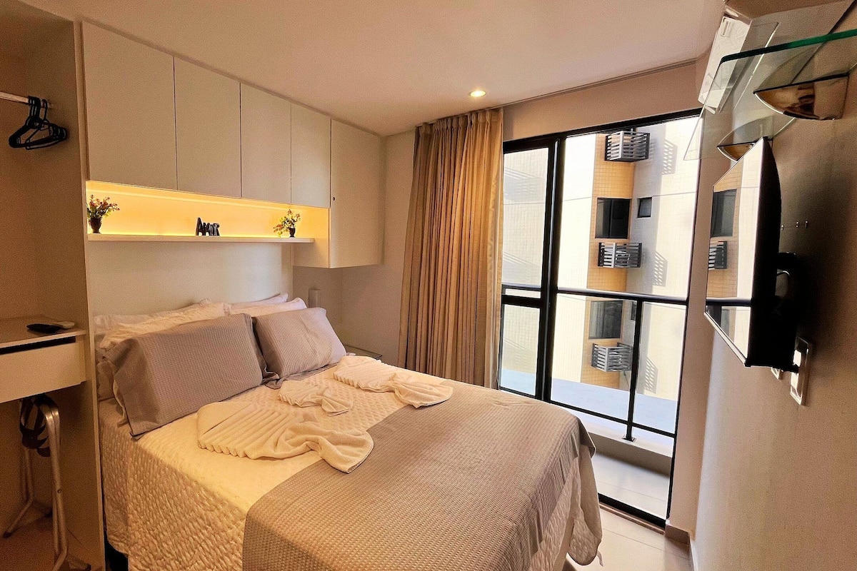Une chambre moderne avec un lit, une armoire, un miroir et une fenêtre donnant sur des immeubles.