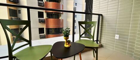 A balcony with two green chairs and a small table, a potted plant, and a view of a multi-story building with balconies.