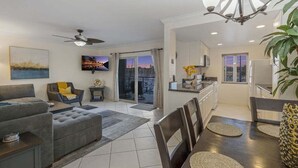 A modern living room with a grey sofa, a dining table with chairs, a ceiling fan, and a kitchen area with cabinets and a countertop. - Serene Harborfront Condo | Unbeatable Views & Coastal Charm (Oceanside)