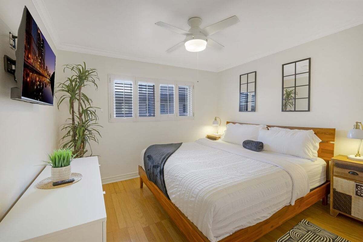 A bedroom with a bed, bedside tables, a ceiling fan, a TV, a window with blinds, and a plant.