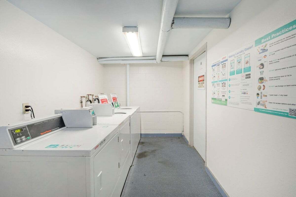 A clean, well-lit laundry room with a washing machine, a dryer, and a wall-mounted ironing station. There are instructional posters on the wall.