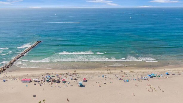 A beach with a long wooden jetty extending into the ocean, sandy beach with people and beach umbrellas, and a clear blue sky. - Serene Harborfront Condo | Unbeatable Views & Coastal Charm (Oceanside)