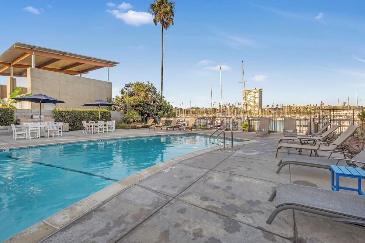 A swimming pool with lounge chairs and umbrellas, surrounded by a concrete deck and palm trees.