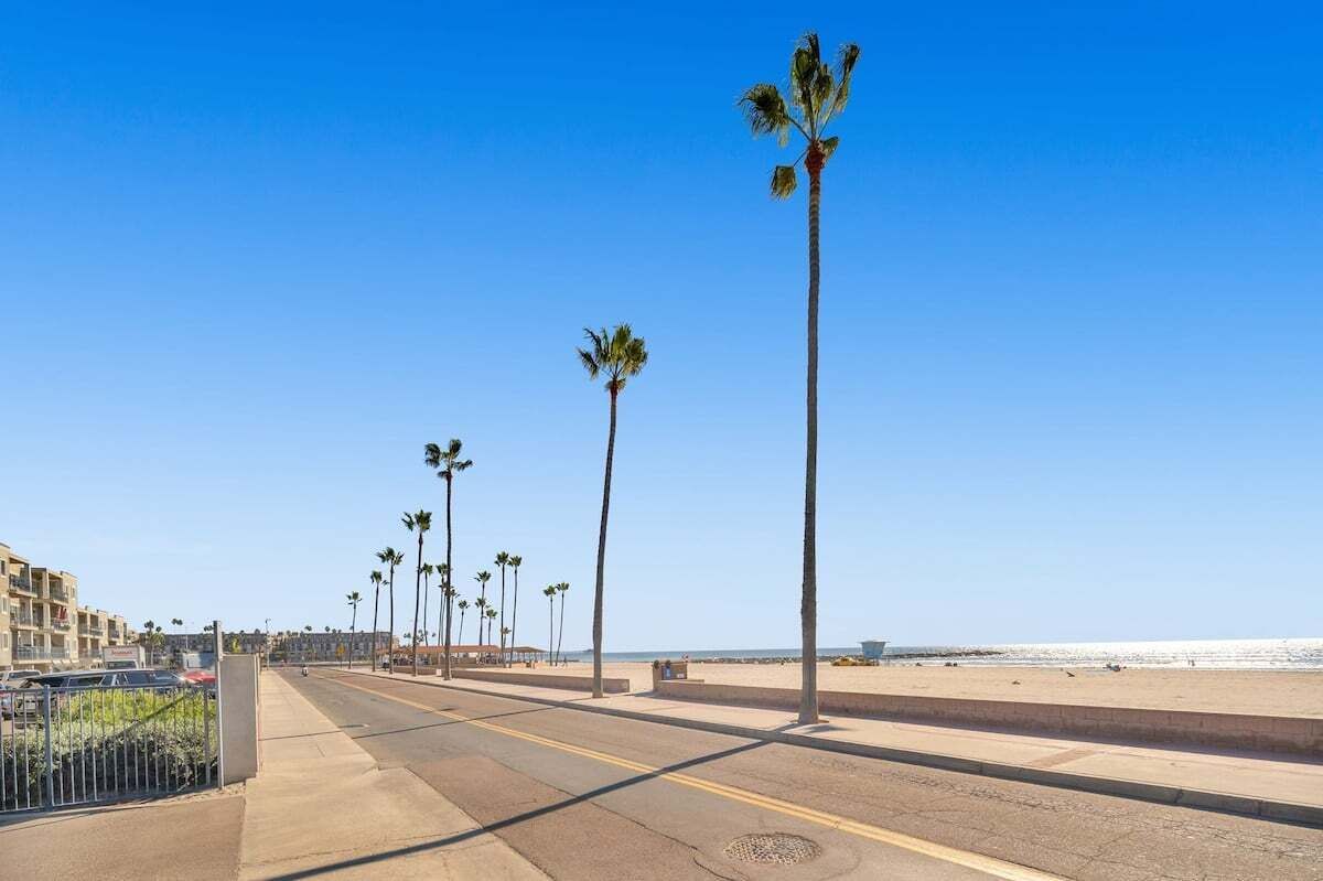 A coastal road lined with palm trees, leading to a beach with a clear sky.