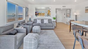Un salon moderne avec un canapé gris, une table à manger avec des chaises et une vue sur la plage grâce à de grandes fenêtres.