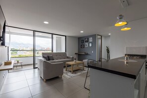 Een modern appartement met een bank, salontafel en eethoek. Het heeft grote ramen, een keuken met fornuis, en uitzicht over de stad.