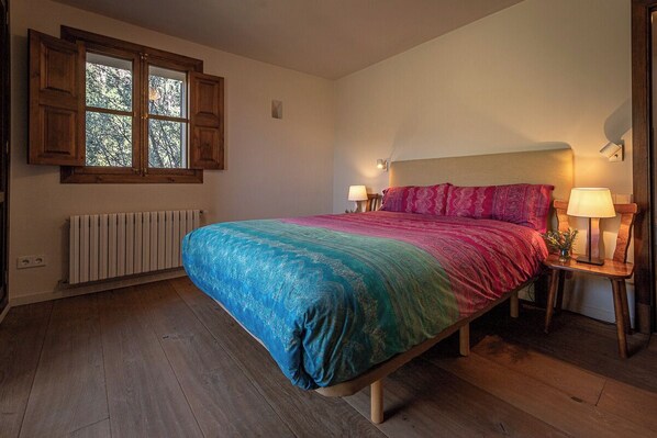 A bedroom with a bed, wooden floor, a radiator, a window with wooden shutters, and two bedside lamps. - Cán Sandy magnificent mountain house with unsurpassed views of the village (Fornalutx)