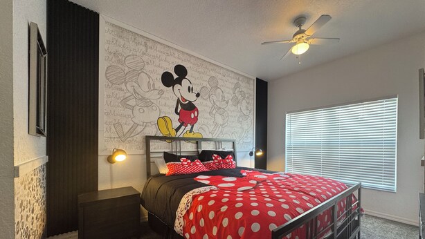 Ein Schlafzimmer mit Wandgestaltung im Mickey-Mouse-Design, einem Bett mit gepunktetem Bettüberwurf, einem Nachttisch, einem Schreibtisch und einem Deckenventilator.