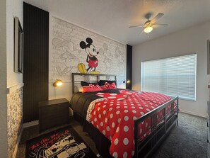 Une chambre avec un mur décoré sur le thème de Mickey Mouse, un lit recouvert d’une couette à pois, une table de chevet, un bureau et un ventilateur de plafond.