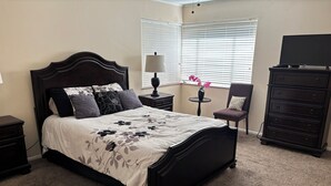 A bedroom with a dark wood bed, a small round table with a lamp, a chair, and a dresser.