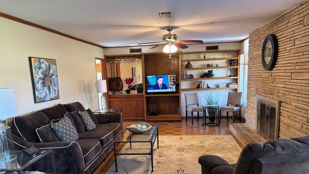 A living room with a sofa, armchair, coffee table, and TV. There is a fireplace, a ceiling fan, and a bookshelf.