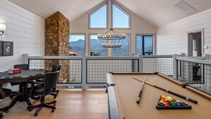 Amplia habitación con mesa de billar, chimenea de piedra y grandes ventanales con vistas a las montañas.