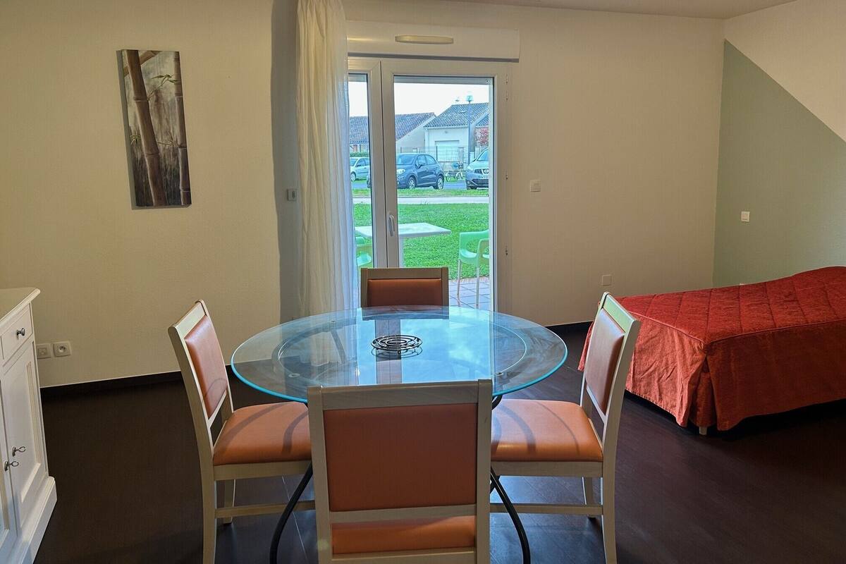 Een kamer met een glazen tafel en stoelen, een bed met een rode deken, een deur naar buiten en een schilderij aan de muur.
