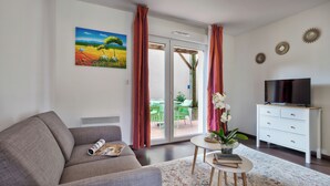 Una sala de estar con un sofá, una mesa de centro con libros y una planta, un televisor en un mueble blanco y un cuadro en la pared.